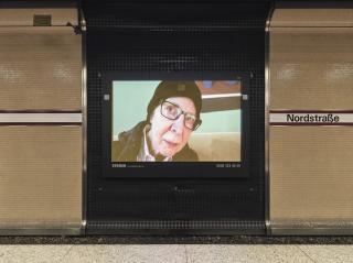 I AM THE FIRST LESBIAN I EVER MET Eine ältere, weiße Person blickt aus einem Werbebildschirm an der U-Bahn-Haltestelle Nordstraße. Ihr Gesicht ist nah an der Kamera und zum Objektiv gewandt. Sie trägt eine dunkel gerahmte Brille, eine schwarze Mütze und eine dicke Jacke, obwohl sie drinnen zu sitzen scheint