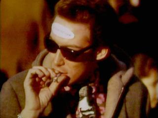 Eine Person mit kurzen, zurückgekämmten Haaren und dunkler Sonnenbrille ist rauchend in Nahaufnahme zu sehen. Auf ihrer Stirn befindet sich ein Protestaufkleber, dessen Text nicht zu erkennen ist. Der Bildhintergrund ist unscharf. Die Person trägt einen Wintermantel und eine Krawatte in der Mode der 1960er-Jahre. Die Farbgebung ist in warmen Tönen, bräunlich bis orange.