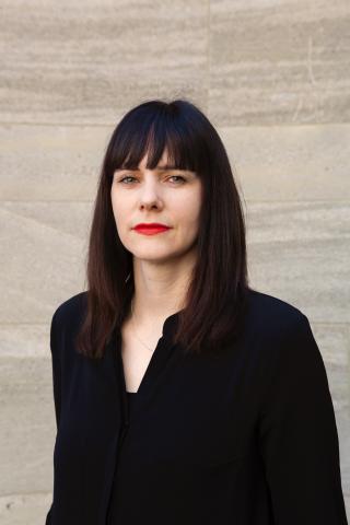 Ein Portrait von Monika Kerkmann mit langen dunklen Haaren und rotem Lippenstift. Sie blickt direkt in die Kamera blickt und trägt eine schwarze Bluse.