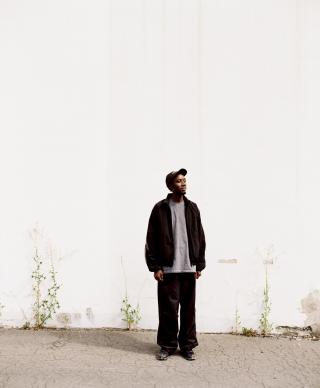 Der Künstler KMRU steht im Außenraum vor einer Wand. Der Boden unter ihm ist rissig, und an der Wand brechen vereinzelt Pflanzen durch die Oberfläche. Der Künstler trägt locker sitzende dunkle Kleidung, ein graues T-Shirt und eine dunkle Cap.