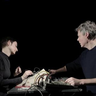 Two musicians sit at a table facing each other. They are brightly lit against a pitch dark background. One is patching a cable on their analoge synthesizer, the other is turning a knob on their mixing board.he musicians are brightly lit against a pitch dark background.