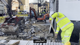 Auf einer Baustelle ist der Gehweg großflächig aufgebrochen. Zwischen Schutthaufen arbeiten mehrere Personen in Warnkleidung. Links bewegt ein großer Baggerarm schwere Betonstücke, weiter hinten bedient jemand einen kleinen Minibagger mit Presslufthammer. Rechts steht eine Person nahe einer Hauswand und stützt sich auf eine Schaufel. Fahrzeuge und Absperrgitter rahmen die Szene in einer belebten Straßenumgebung. Am unteren Bildrand steht eine Bildunterschrift mit der Aufschrift „…ja/nein?…“.