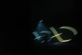 Ein dunkler Raum wird punktuell durch einen Scheinwerfer erhellt. Im Licht dieses Scheinwerfers bewegen sich abstrakte Formen in türkis und gelb.