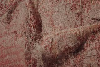 Nahaufnahme eines grob gewebten beige-roten Tuchs. Die Textilstruktur ist an vielen Stellen abgenutzt und zerschlissen, die rote Farbe ist ausgeblichen und bröckelt.