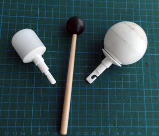 Zwei Spitzen eines Blindenlangstocks (Zylinder und Kugel) liegen auf einer Unterlage mit Gitterlinien. Zwischen den Spitzen liegt der Schlägel eines Schlaginstruments.