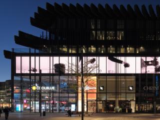 A popular shopping street in Düsseldorf is nearly empty, late in the evening. The stores in the large Kö-Bögen II complex are dimly lit. Above them stretches a 120-meter-long LED wall displaying a video work by Lucas Aal that explores the destruction of a camera.