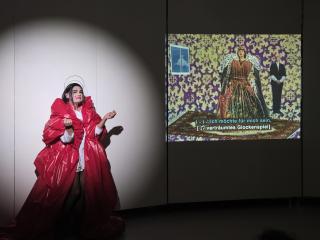 Rafael-Evitan Grombelka steht in einem voluminösen roten Kostüm neben der Projektion von Akiko Hadas „The Fall of a Queen, or The Taste of the Fruit to Come“ (1991). Die Projektion zeigt eine Königin und ihren königlichen Begleiter.
