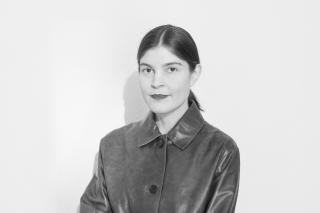 Schwarz-Weiß-Porträt von Judith Greitemann. Die Person trägt eine dunkle Lederjacke, blickt direkt in die Kamera und steht vor einer hellen Wand.