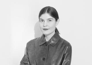 lack-and-white portrait of Judith Greitemann with her dark hair pulled back and wearing a dark leather coat. She looks directly into the camera against a white background.