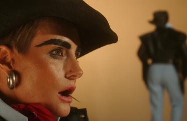Ein Gesicht im Profil mit Cowboyhut, Ohrringen, gemalten Augenbrauen und offenem Mund mit einem Zahnstocher. Im Hintergrund die Silhouette eines Cowboys von hinten.