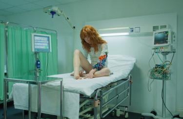 Das Bild zeigt eine junge rothaarige Frau, die auf einem Bett in einem Krankenhaus sitzt.