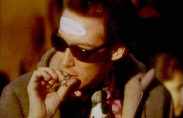 Eine Person mit kurzen, zurückgekämmten Haaren und dunkler Sonnenbrille ist rauchend in Nahaufnahme zu sehen. Auf ihrer Stirn befindet sich ein Protestaufkleber, dessen Text nicht zu erkennen ist. Der Bildhintergrund ist unscharf. Die Person trägt einen Wintermantel und eine Krawatte in der Mode der 1960er-Jahre. Die Farbgebung ist in warmen Tönen, bräunlich bis orange.