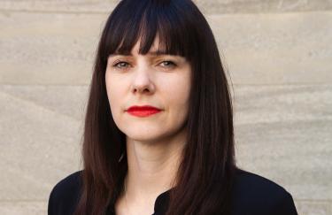 Ein Portrait von Monika Kerkmann mit langen dunklen Haaren und rotem Lippenstift. Sie blickt direkt in die Kamera blickt und trägt eine schwarze Bluse.