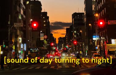 Eine urbane Straße in der Dämmerung. Links und rechts umgeben von hohen Gebäuden. Mehrere rote Ampeln und leuchtende Straßenlaternen ziehen sich entlang der Straße, dazu die Scheinwerfer vorbeifahrender Autos. Unten leuchtet der Himmel in Orange, während er nach oben hin ins Blau übergeht. Am unteren Bildrand erscheint in gelber Schrift die Texteinblendung [sound of day turning to night].