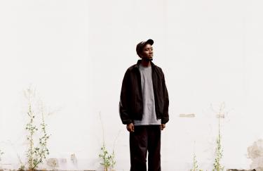 Der Künstler KMRU steht im Außenraum vor einer Wand. Der Boden unter ihm ist rissig, und an der Wand brechen vereinzelt Pflanzen durch die Oberfläche. Der Künstler trägt locker sitzende dunkle Kleidung, ein graues T-Shirt und eine dunkle Cap.