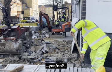 Auf einer Baustelle ist der Gehweg großflächig aufgebrochen. Zwischen Schutthaufen arbeiten mehrere Personen in Warnkleidung. Links bewegt ein großer Baggerarm schwere Betonstücke, weiter hinten bedient jemand einen kleinen Minibagger mit Presslufthammer. Rechts steht eine Person nahe einer Hauswand und stützt sich auf eine Schaufel. Fahrzeuge und Absperrgitter rahmen die Szene in einer belebten Straßenumgebung. Am unteren Bildrand steht eine Bildunterschrift mit der Aufschrift „…ja/nein?…“.