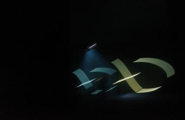 Ein dunkler Raum wird punktuell durch einen Scheinwerfer erhellt. Im Licht dieses Scheinwerfers bewegen sich abstrakte Formen in türkis und gelb.