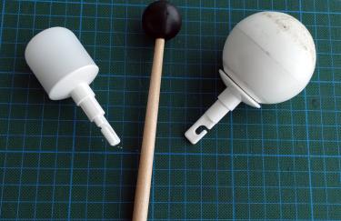 Zwei Spitzen eines Blindenlangstocks (Zylinder und Kugel) liegen auf einer Unterlage mit Gitterlinien. Zwischen den Spitzen liegt der Schlägel eines Schlaginstruments.