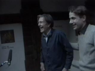 John Cage: Mushroom et Variationes, 1985. Videostill Zwei Männer im Profil lachen, links ist John Cage, rechts ist Axel Wirths, der John Cage dabei den Arm auf den Rücken legt.