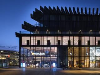 Das Foto zeigt ein modernes, mehrstöckiges Gebäude bei Nacht. Die Fassade besteht größtenteils aus Glas und gibt den Blick in ein hell beleuchtetes Erdgeschoss frei, in dem ein Geschäft mit „Coolblue“-Schriftzug zu sehen ist. Über dem transparenten Sockel erhebt sich ein dunkler, markanter Aufbau mit vertikalen Lamellen und begrünten Elementen. Der Vorplatz ist weitläufig und ruhig, nur schwach beleuchtet, wodurch die Architektur besonders hervorgehoben wird.