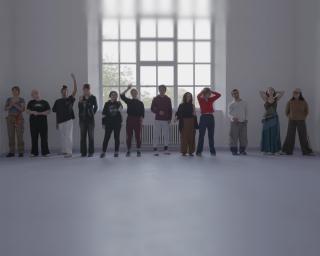 Eine Gruppe von zwölf jungen Menschen steht nebeneinander in einem hellen, minimalistischen Raum. Hinter ihnen befindet sich ein großes Fenster, durch das Tageslicht fällt, dessen oberer Bereich sanft in Unschärfe übergeht. Die Personen tragen unterschiedliche Kleidung und nehmen verschiedene Körperhaltungen ein, einige mit Armen nach oben gestreckt, andere mit verschränkten Händen.