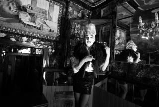 Schwarz-weißes Reenactment einer Karl Lagerfeld-Serie aus den 1980er-Jahren im Berliner Club Kumpelnest 3000. Rafael-Evitan Grombelka übernimmt die Rolle von Gunter Trube und reinszeniert Look und Pose direkt im Originalraum. Mit hochgestecktem Haar, Make-Up, kurzem Rock, Bluse und federartiger Stola nimmt er die Bildsprache der Serie auf. Spiegel, gemusterte Wände und ein Kronleuchter gliedern den Raum, im Hintergrund ist ein Wandbild zu sehen, das ein Kind zeigt, das an einen Löwen angelehnt ist.