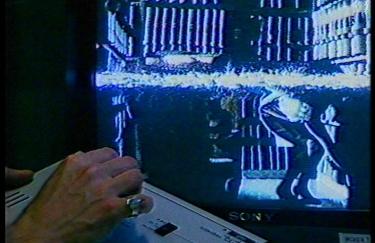 Eine Hand bedient einen Joystick vor einem Bildschirm. Auf dem Bildschirm ist eine Person in Rock und Stöckelschuhen zu erkennen, die sich bückt, um einen großen Aktenordner aus einem Regal voller Dokumente zu holen.