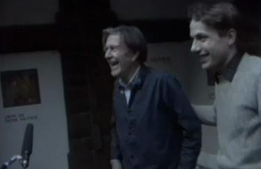 Zwei Männer im Profil lachen, links ist John Cage, rechts ist Axel Wirths, der John Cage dabei den Arm auf den Rücken legt.