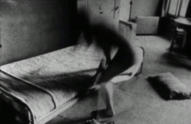 Black-and white image of a blurry figure changing in front of a one-person bed. Film still from Hooykaas/Stansfield, Daydreams, 1972.