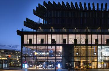 Das Foto zeigt ein modernes, mehrstöckiges Gebäude bei Nacht. Die Fassade besteht größtenteils aus Glas und gibt den Blick in ein hell beleuchtetes Erdgeschoss frei, in dem ein Geschäft mit „Coolblue“-Schriftzug zu sehen ist. Über dem transparenten Sockel erhebt sich ein dunkler, markanter Aufbau mit vertikalen Lamellen und begrünten Elementen. Der Vorplatz ist weitläufig und ruhig, nur schwach beleuchtet, wodurch die Architektur besonders hervorgehoben wird.