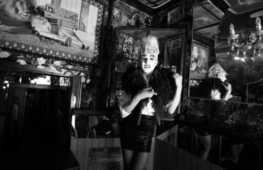 Schwarz-weißes Reenactment einer Karl Lagerfeld-Serie aus den 1980er-Jahren im Berliner Club Kumpelnest 3000. Rafael-Evitan Grombelka übernimmt die Rolle von Gunter Trube und reinszeniert Look und Pose direkt im Originalraum. Mit hochgestecktem Haar, Make-Up, kurzem Rock, Bluse und federartiger Stola nimmt er die Bildsprache der Serie auf. Spiegel, gemusterte Wände und ein Kronleuchter gliedern den Raum, im Hintergrund ist ein Wandbild zu sehen, das ein Kind zeigt, das an einen Löwen angelehnt ist.