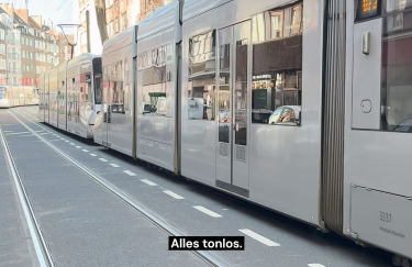 Ein moderner, silberner Straßenbahnzug fährt durch eine städtische Straße mit mehrstöckigen Wohnhäusern im Hintergrund. Die Gleise verlaufen parallel zur Fahrbahn, während Autos am Straßenrand parken. Unten im Bild ist der Untertitel „Alles tonlos.“ eingeblendet.