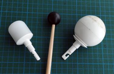 Zwei Spitzen eines Blindenlangstocks (Zylinder und Kugel) liegen auf einer Unterlage mit Gitterlinien. Zwischen den Spitzen liegt der Schlägel eines Schlaginstruments.