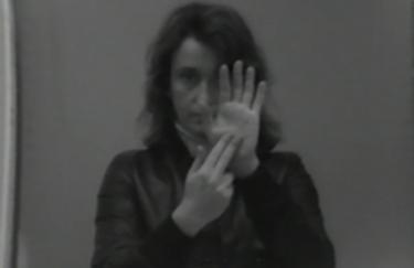 Ein schwarz-weißes Videostill der Künstlerin VALIE EXPORT. Die Künstlerin blickt in die Kamera und formt ein Zeichen mit den Händen. Es ist das Zeichen für den Buchstaben „n“ im Zweihand-Fingeralphabet. Intendiert ist aber vermutlich der Buchstabe „m“.