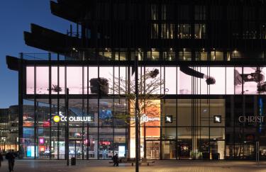 Eine beliebte Einkaufsstraße in Düsseldorf ist zur späten Abendstunde nahezu leer. Die Geschäfte in dem großen Gebäudekomplex Kö-Bögen II sind nur noch schwach beleuchtet. Über ihnen erstreckt sich eine 120 Meter lange LED Wand auf der eine Videoarbeit von Lucas Aal läuft, in der die Zerstörung einer Kamera thematisiert wird.