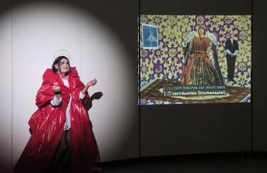 Im Storage Museum führt Rafael-Evitan Grombelka eine Simultanübersetzung von Akiko Hadas Videooper „The Fall of a Queen“ auf. Rafael-Evitan steht im Rampenlicht links neben der untertitelten Videoprojektion. Er ist in einem roten Kostüm von Gunter Trube gekleidet, das fast genauso aussieht, wie die Königin in Akiko Hadas Arbei rechts neben ihm.