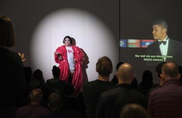 Rafael-Evitan Grombelka steht vor einem großen Publikum im Storage Museum. Im Rampenlicht führt er eine Simultanübersetzung von Akiko Hadas Videooper „The Fall of a Queen“ auf. In dem Video ist ein junger Mann in einem Anzug zu sehen. Die Untertitel lesen „Nur weil sie aus königlichem Geblüt stammt…“. Rafael-Evitan steht verkleidet als die Königin links daneben und reagiert mit einem verwirrten Gesichtsausdruck auf diese Äußerung.