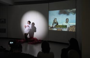 Rafael-Evitan Grombelka performt eine Simultanübersetzung von Akiko Hadas „The Fall of a Queen“. In dem Video tanzen zwei junge Menschen zu den Untertiteln: “Wer möchte nicht auf einem Thron sitzen? [unheilvolle Orgeln]”. Rafael-Evitan steht links daneben und tanzt genauso wie die zwei im Video.
