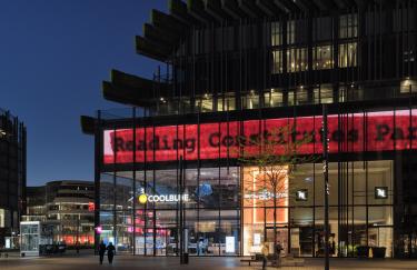 Spät am Abend ist eine beliebte Einkaufsstraße in Düsseldorf nahezu leer. Die Geschäfte in dem großen Gebäudekomplex Kö-Bögen II sind nur noch schwach beleuchtet. Über ihnen erstreckt sich eine 120 Meter lange LED Wand auf der eine Videoarbeit von Hu Xinyue läuft und ein Blatt Papier zeigt, auf dem “Reading Constitutes Participation” in schwarzer Druckschrift und roter Markierung steht.