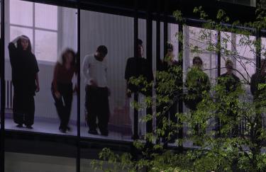 Ein großer LED Screen zeigt die Videoarbeit von Lucien Liebecke, in der mehrere Studierende nebeneinander in einer Reihe stehen und unterschiedliche Bewegungen ausführen. Das Video ist teilweise von dünnen vertikalen Stahlträgern des Gebäudes verdeckt, die die dargestellte Personengruppe rhythmisch untergliedert. Außerdem sind Zweige und grüne Blätter eines Baumes rechts im Vordergrund.