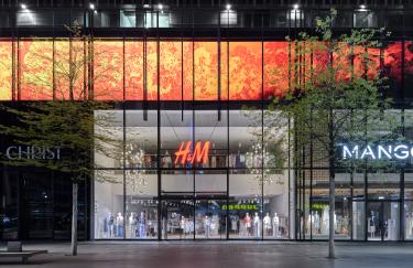 Eine Frontalansicht von drei Modegeschäften auf der Schadowstraße, die zur späten Abendstunde geschlossen sind. Über ihnen leuchtet eine große LED-Wand in den lebhaften Orange- und Rottönen eines Videokunstwerks von Ole Christian Dreihaupt.