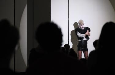 Rafael-Evitan Grombelka performt Deaf Cinderella vor einem großen Publikum im Storage Museum. Er trägt eine blonde Perrücke, eine weiße Bluse mit schwarzer Fellweste, einen schwarzen Paillettenrock mit Strumpfhose und weißes Makeup mit rosen Lippenstift.