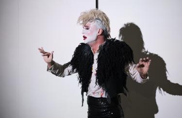 Rafael-Evitan Grombelka während seiner Deaf Cinderella Performance. Er wird von einem weißen Scheinwerferlicht angestrahlt und wirft daher einen Schatten hinter sich. Mit den Händen gebärdet er die Wörter, die man ihm nicht von seinem emotionalen Gesichtsausdruck ablesen kann.