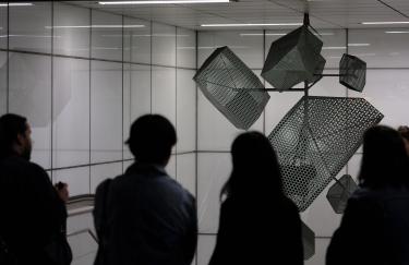 Eine futuristisch anmutende Klanginstallation hängt von der Decke des weiß gefliesten Modellraums im U-Bahnhof Heinrich-Heine-Allee. Davor stehen Menschen, die sich KMRUs Audio Stück anhören.