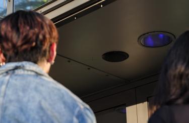Ein Blick nach oben zur Decke des Notausgangs des UFA Palast Düsseldorfs.In einer Metallplatte sind ein Lautsprecher und ein blau leuchtendes Licht eingebaut. Im Vordergrund stehen zwei Menschen, die ihre Köpfe in diese Richtung gedreht haben, um KMRUs FOLD II zu hören.