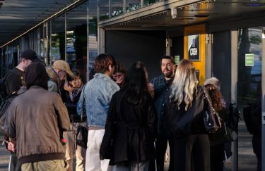 Joy Bausch steht vor dem Notausgang des UFA Palast Düsseldorfs mit einer Führungsgruppe. In der Decke über ihm ist ein Lautsprecher der KMRUs Audiostück abspielt.