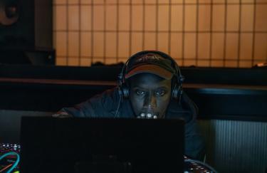 In einem dunklen Raum sitzt KMRU mit Kopfhörern hinter seinem Laptop und Mischpulten. Mit seiner rechten Hand steuert er seinen Computer, während seine linke Hand auf seinen Kopf stützt. Hinter ihm sind ein großer Lautsprecher und helle Wandfliesen zu sehen.