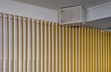 In der oberen Ecke des Salon des Amateurs hängt großer, weißer Lautsprecher von dem KMRUs FOLD II abgespielt wird. Die Wand unterhalb des Lautsprechers ist mit einer Vielzahl weiß-gelber Wandpaneele verkleidet, die vertikale Streifen erzeugen.