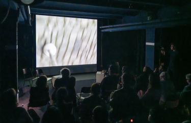 Im Keller der Filmwerkstatt wird Dirk Sorges „Frotter la Cité“ Video vor einem sitzenden Publikum abgespielt. In erster Reihe sitzt Sorge mit einem Mikrofon, um zu beschreiben, was gerade zu sehen ist: ein runder Kopf eines Blindenstocks auf einem gepflasterten Weg.
