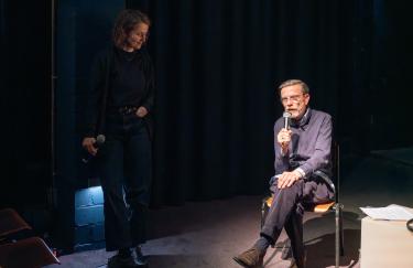 Marcel Odenbach sitzt auf einem Stuhl auf der Bühne der Filmwerkstatt und erzählt über sein ausgestelltes Werk. Links neben ihm steht Linnea Semmerling und hört ihm gespannt zu.